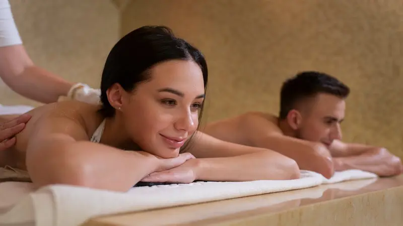 A woman resting her chin on her hands and a man behind her, both lying face down on side-by-side treatment tables in a warm stone-walled room, with a therapist's hands working across the woman's shoulder.