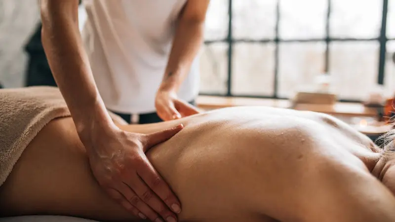 A therapist in a white top pressing both hands into a client's lower back on a massage table, with soft daylight coming through a large window behind them.