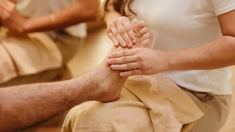 A seated guest's bare foot resting on a folded towel across a therapist's lap, the therapist's thumbs pressing along the top of the foot, in a warm cream-toned room.