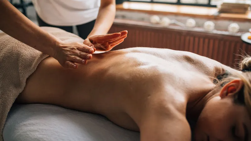 A therapist in a white top working both hands along the lower back of a woman lying face down on a massage table, draped with a beige towel, beside a bright window lined with candles.
