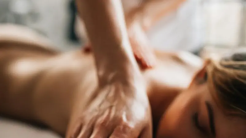 A therapist's forearm and hand pressing along the shoulder and upper back of a woman lying face down on a massage table with her eyes closed, in a warm, softly lit room.