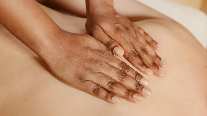 Both of a therapist's hands resting flat across a client's upper back on a massage table, in a room lit in warm cream tones.