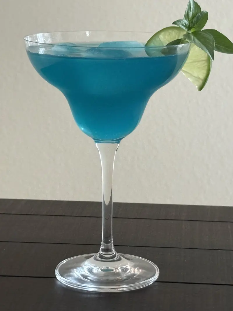 Blue Hawaiian drink in a coupe glass, bright blue, garnished with a lime wheel and mint.