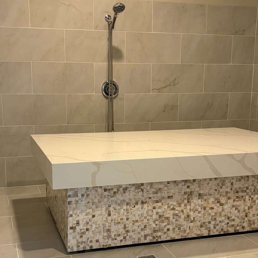 The Moroccan bath room at Healing Spa — a heated white marbled stone table on a mother-of-pearl mosaic base, standing in a marble-tiled wet room with a handheld shower on the wall behind it and a floor drain alongside