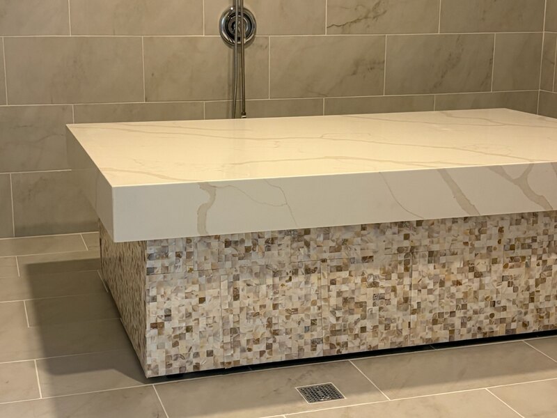 The hammam room at Healing Spa — a heated white marbled stone table on a mother-of-pearl mosaic base, standing in a marble-tiled wet room with a handheld shower on the wall behind it and a floor drain alongside